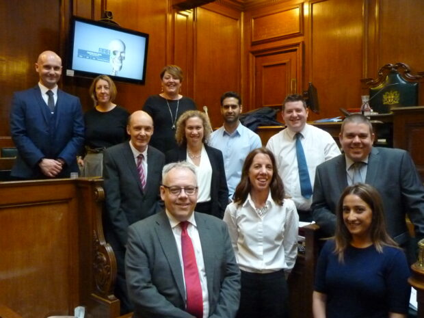 Group of people standing in a wood-cladded, old-fashioned courtroom, posing for the camera. There's a Clickshare monitor in the room.